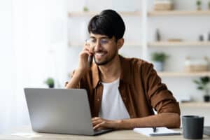 Cheerful indian guy businessman sitting at workdesk with laptop having phone conversation with business partner or client, typing on computer keyboard, office interior, copy space